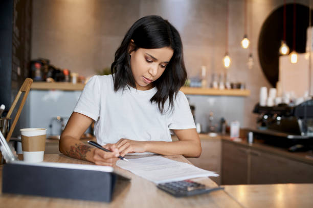 mujer dueña de un café firmando papeles calculando gastos comerciales - loan fotografías e imágenes de stock