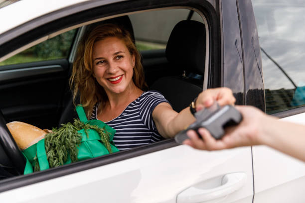 Paying for her curbside pickup with a credit card Selective focus shot of cheerful mid adult woman sitting in her car, in a drivers seat and playing for groceries with a credit card during a curbside pickup. curbsidepickup stock pictures, royalty-free photos & images