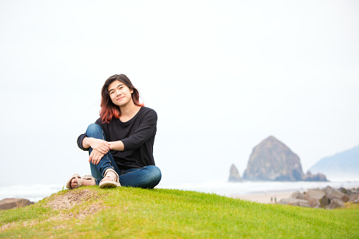Biracial teen girl sitting on grassy knoll or hill by shore near the ocean in the Pacific Northwest on foggy day