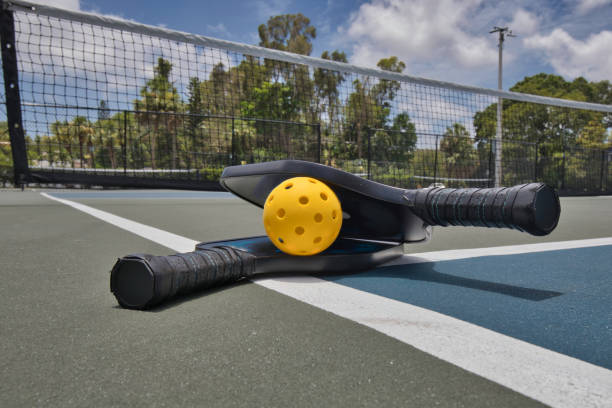 Pickleball and Paddles Pickleball and paddles laying on a court in front of a net on a sunny day. pickleball stock pictures, royalty-free photos & images