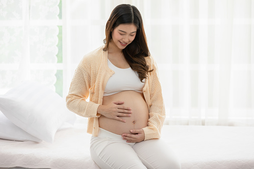Happy Pregnant Woman sitting on bed holding and stroking her big belly at home,Pregnancy of young woman enjoying with future life relax at home,Motherhood and Pregnant Concept,Soft focus