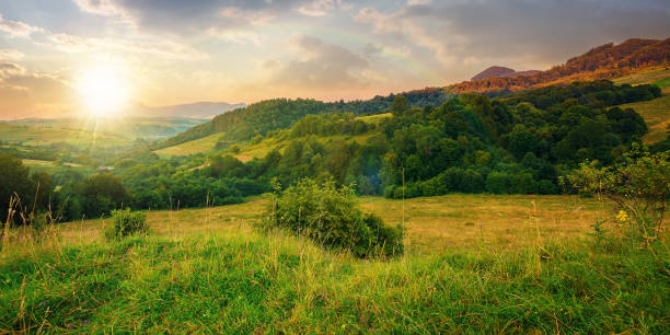 mountainous rural landscape at sunset. beautiful scenery with forests, hills and meadows in evening light. ridge with high peak in the distance. village in the distant valley