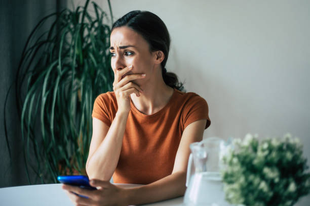 悲しい、イライラした若いブルネットの女性は、彼女がアパートの椅子に座っている間、手にスマートフォンで泣いています - 半狂乱 ストックフォ��トと画像