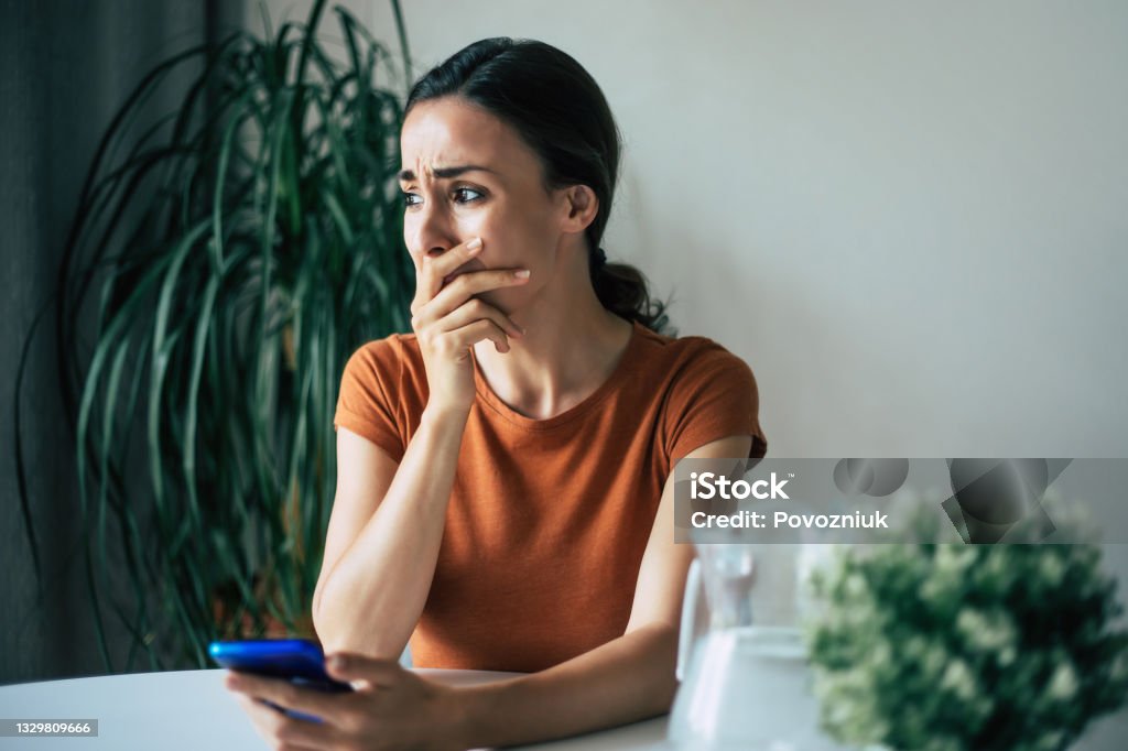 Sad, frustrated young brunette woman is crying with smartphone in hands while she sitting on the chair at apartment Women Stock Photo