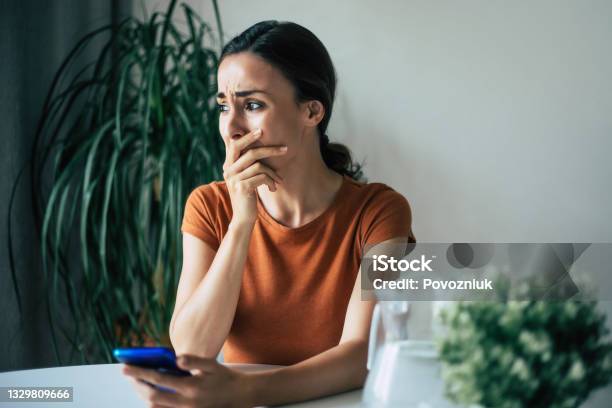 Sad Frustrated Young Brunette Woman Is Crying With Smartphone In Hands While She Sitting On The Chair At Apartment Stock Photo - Download Image Now