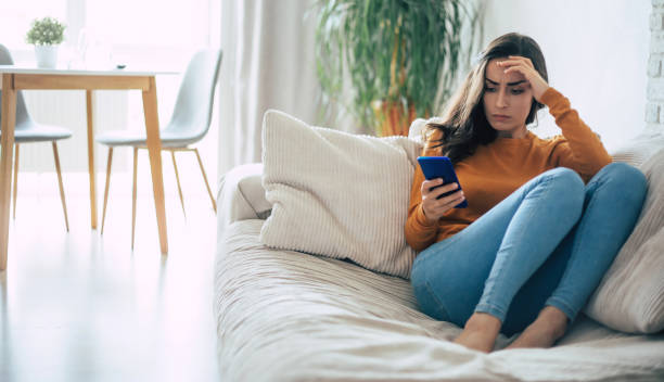 istock Frustrated and depressed young brunette woman is crying with a smartphone in hands while she sitting on the couch at home 1329809687