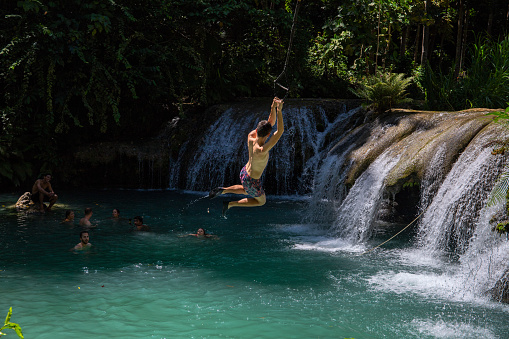 Siquijor, the Philippines - 12 Mar 2020: Boy jumping with tarzan rope over waterfall. Rope swing jump into waterfall. South Asia ecotourism and adventure. Young traveler on gap year. Extreme sport