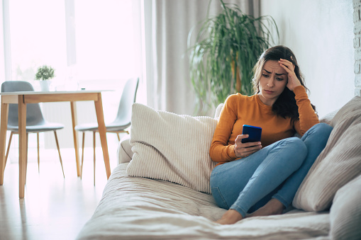 Frustrated and depressed young brunette woman is crying with a smartphone in hands while she sitting on the couch at home
