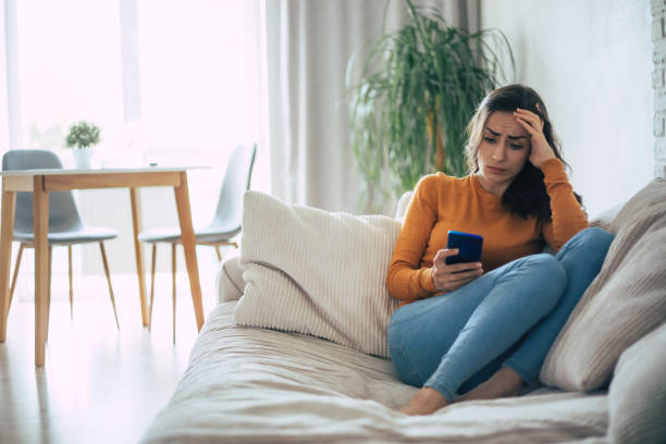 Frustrated and depressed young brunette woman is crying with a smartphone in hands while she sitting on the couch at home stock photo