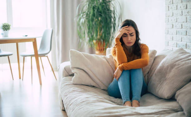 istock Young sad Brunette woman with severe headache holds her head while she sitting on the couch at home 1329809765