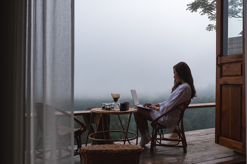 A young asian woman using and working on laptop computer while sitting on balcony with a beautiful nature view on foggy day