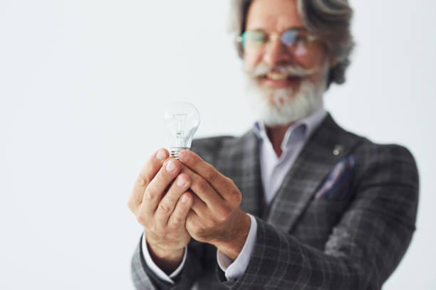 istock Posing for a camera while standing against white background. Senior stylish modern man with grey hair and beard indoors 1329837043