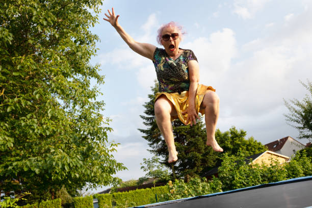 Eccentric senior woman with dyed pink hair, 67 years old, wearing sunglasses, a golden skirt and black shirt with colorful pattern, jumping on a trampoline in backyard, outdoors at a summer evening, she has overweight and has fun exercising