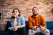 Front view of happy cheerful young couple holding controllers and playing video games on console sitting together on couch