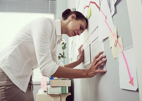 Female office worker bent over with head lening to a wall and negative business chart.