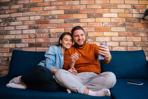 Middle shot portrait of happy young couple having online webcam chat with via video call on mobile phone, sitting together on comfortable soft sofa at cozy living room. Concept of household life.