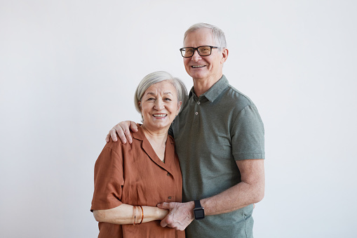 Waist up portrait of happy senior couple embracing and looking at camera while standing against white background, copy space