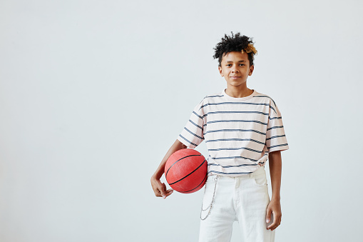 Minimal portrait of mixed-race teenage boy holding basketball ball while standing against white background, copy space