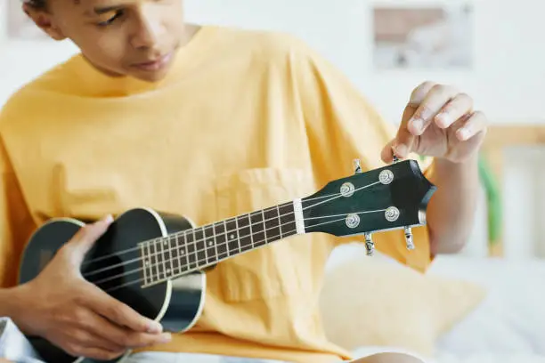 Minimal cropped shot of teenage boy playing ukulele while sitting on bed, copy space