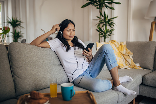 One woman, beautiful Asian woman sitting on the sofa in living room at home. She is using smart phone and listening music on headphones.