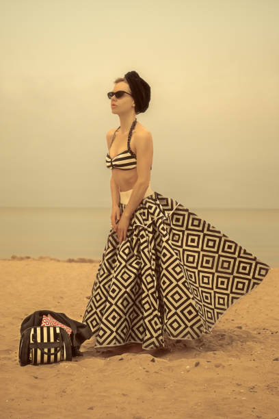 Young vintage woman wearing a parasol as a skirt, sunglasses, and a towel wrapped around her head, standing on the beach with her bathing bag, in front of the ocean The skirt was created with a parasol, designed by the model and stylist of the photography duo. beach umbrella photos stock pictures, royalty-free photos & images