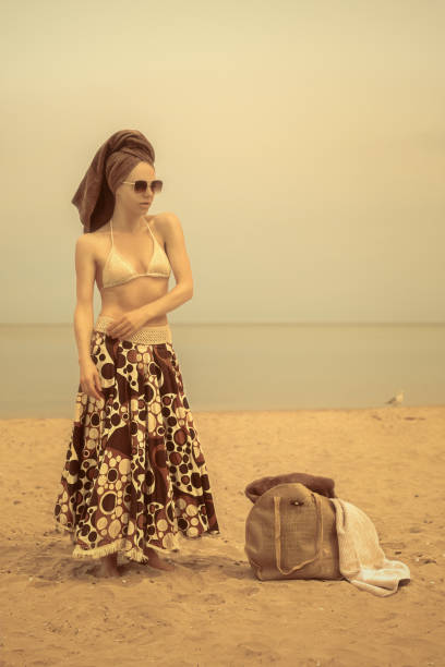 young vintage woman wearing a parasol as a skirt, sunglasses, and a towel wrapped around her head, standing on the beach with her bathing bag, in front of the ocean - image created 1960s 1960s style beach women imagens e fotografias de stock