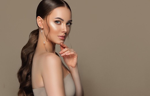 Perfect looking gorgeous young woman wearing in splendid makeup and long ,thin earrings with brown hair gathered in a dense, long ponytail. Straight look of beautiful blue eyes. Beauty, makeup and hairstyle.