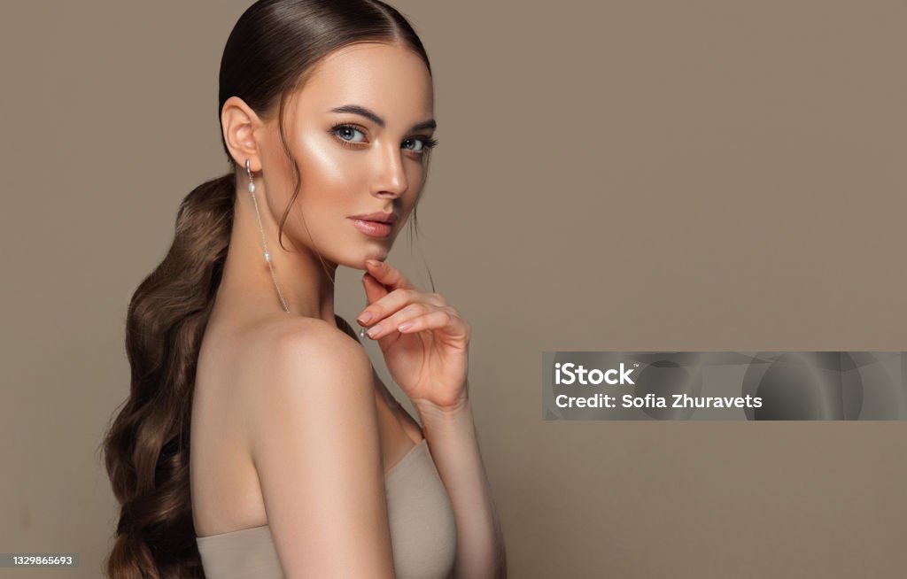 Portrait of perfectly looking young brown haired woman with long hair, wearing in exquisite makeup. Hairstyle and makeup. Perfect looking gorgeous young woman wearing in splendid makeup and long ,thin earrings with brown hair gathered in a dense, long ponytail. Straight look of beautiful blue eyes. Beauty, makeup and hairstyle. Ponytail Stock Photo