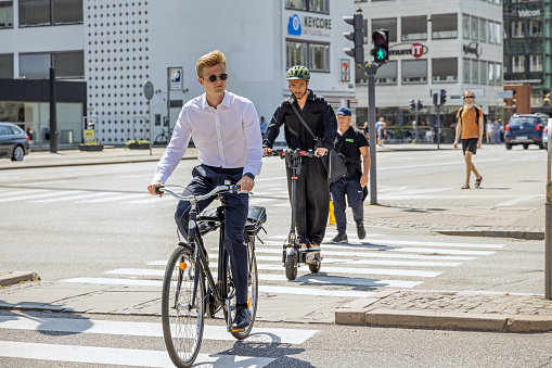 Young men crossing the road on bike and an electrical push scooter on a hot summers day in central Copenhagen