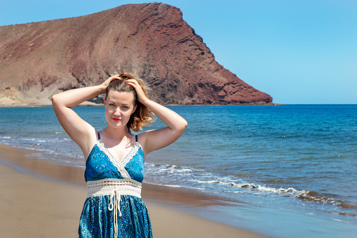 Beautiful Caucasian millennial woman at Playa La Tejita in a sunny summer day with miles of clean golden sand behind and the volcanic Montana Roja in the background in Tenerife, Canary Islands, Spain.