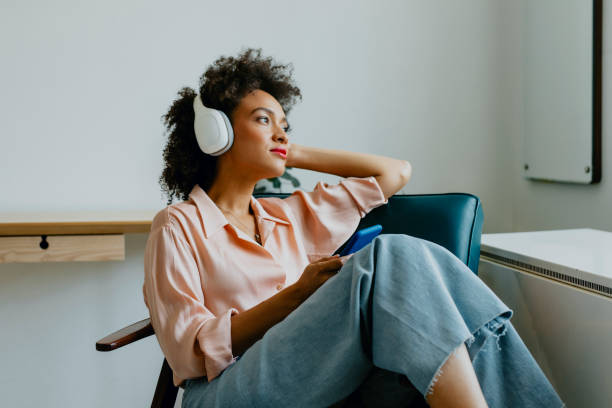 giovane donna rilassata con le cuffie, seduta su una poltrona e ascoltando il suo podcast preferito - udire foto e immagini stock