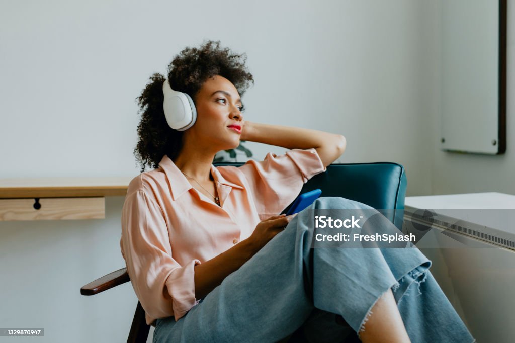 Relaxed Young Woman with Headphones on, Sitting in an Armchair and Listening to her Favorite Podcast A smiling Afro woman sitting comfortably in an armchair with her earphones on, listening to music on her smartphone Podcasting Stock Photo