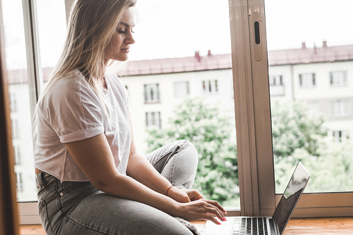 Young woman dressed casually working on laptop while sitting on the window sill at home. Work from home at cozy atmosphere concept
