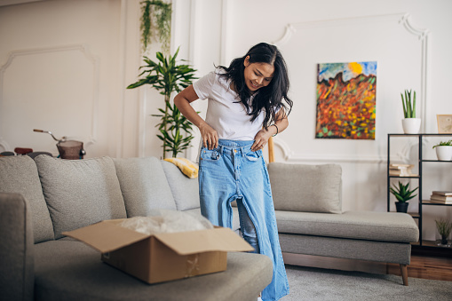 One woman, beautiful Asian woman standing in living room at home. She is trying on denim pants that she ordered.