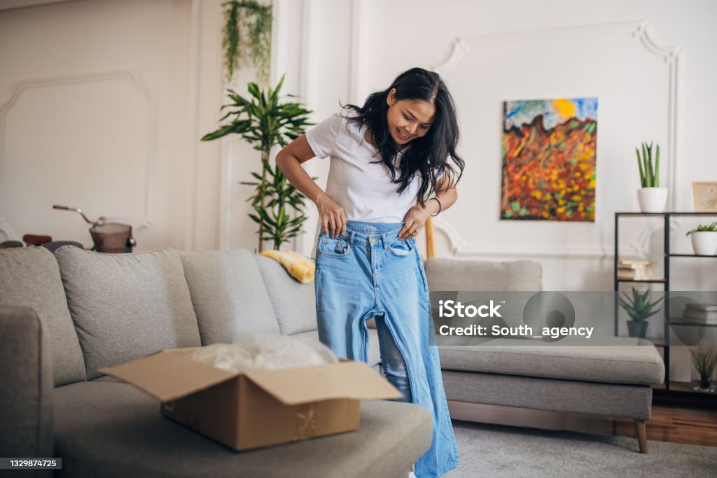 Woman received a package at home One woman, beautiful Asian woman standing in living room at home. She is trying on denim pants that she ordered. Trying On Stock Photo