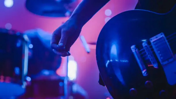 Photo of Rock Band Performing at a Concert in a Night Club. Close Up Shot of a Musician Holding and Dropping a Guitar Pick. Live Music Party in Front of Bright Colorful Strobing Lights on Stage.