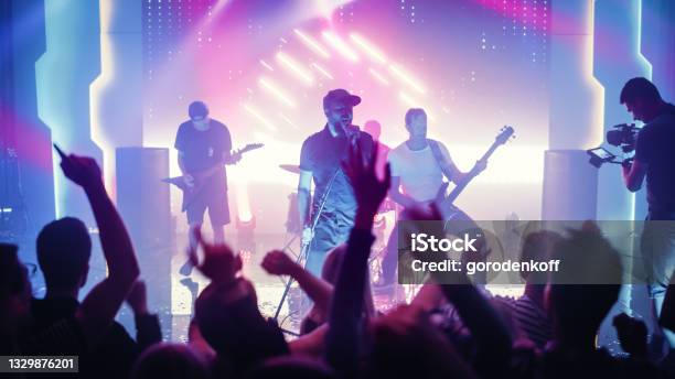 Rock Band With Guitarists And Drummer Performing At A Concert In A Night Club Front Row Crowd Is Partying Silhouettes Of Fans Raise Hands In Front Of Bright Colorful Strobing Lights On Stage Stock Photo - Download Image Now