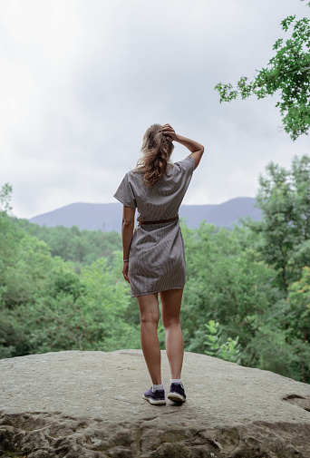Travel and freedom concept. Back view of a young woman in summer dress standing on a big rock in the forest looking away