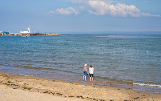 Scarborough, UK. July 9, 2021.  Two women paddle in the sea carrying their shoes in their hands.  A lighthouse and pier is in the distance and a boat is on the water