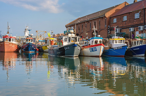 Scarborough, UK. July 9, 2021.  Scarborough, UK. July 9, 2021.  The harbour at Scarborough.  Fishing boats are moored in the harbour alongside a wharf with red-brick buildings.  A cloudy sky is above.