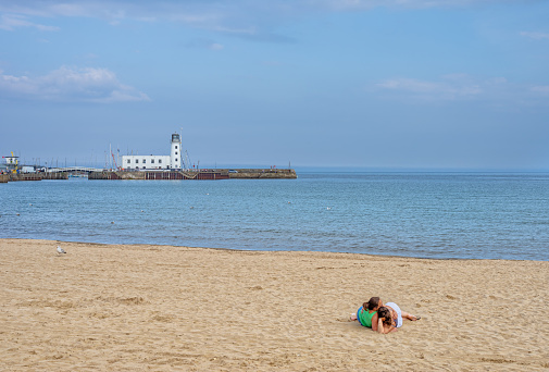 Scarborough, UK. July 9, 2021.  Two people relax close together on a beach in summer. A lighthouse and pier is in the distance and a sky with clouds is  is overhead.