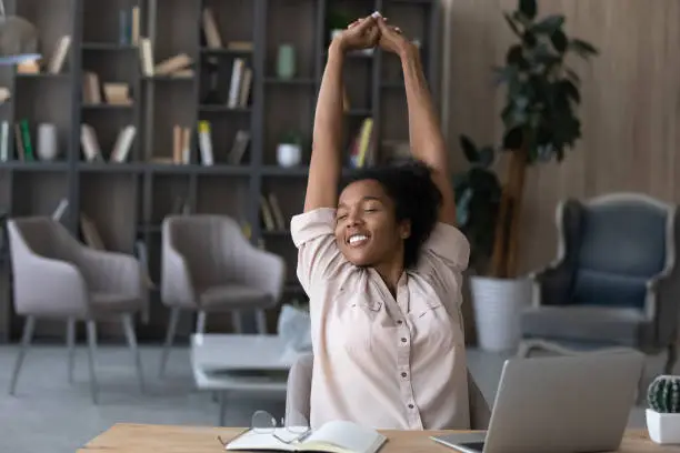 Happy carefree African American woman stretching hands at workplace, leaning back in comfortable chair, student or freelancer relaxing enjoying break after work done, sitting at desk with laptop