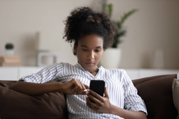 Focused African American woman using smartphone, sitting on couch Focused African American young woman using smartphone, sitting on couch, confident young female looking at phone screen, typing writing message in social network, browsing apps, chatting online scrolling stock pictures, royalty-free photos & images