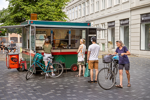 An elderly woman walking her bike in front of a traditionally Danish street food stall in the pedestrian zone in central Copenhagen