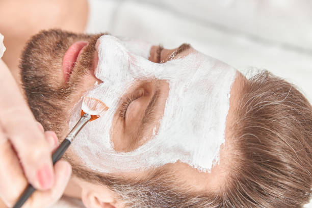 istock Close up bearded young man is relaxing while cosmetologist is spreading white clay on his face 1329886870