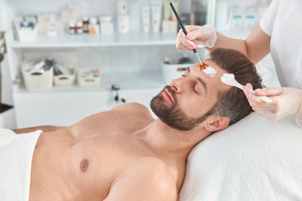 istock Bearded young man is relaxing while cosmetologist is spreading white clay on his face with brush at beauty salon 1329886874