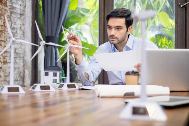 istock Architect sitting in front of computer Holding a blueprint and pointing a pencil at the windmill on the desk in the office. 1329893025