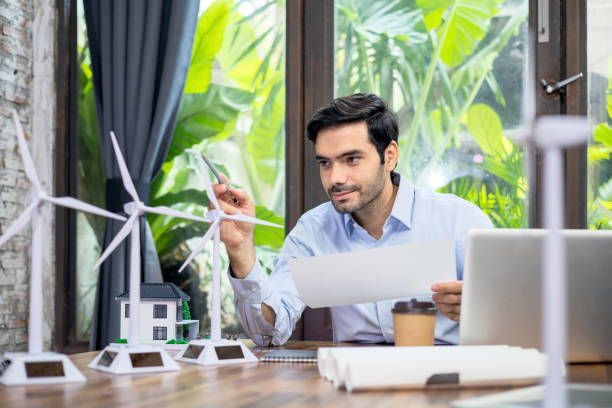 istock Engineer sitting in front of computer Holding a blueprint and pointing a pencil at the windmill on the desk in the office. 1329893044