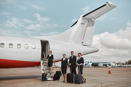 Flight attendant and stewardesses on runway and ladder near airplane jet. Modern passenger plane. Man and women with baggage wear uniform. Teamwork. Civil commercial aviation. Air travel concept