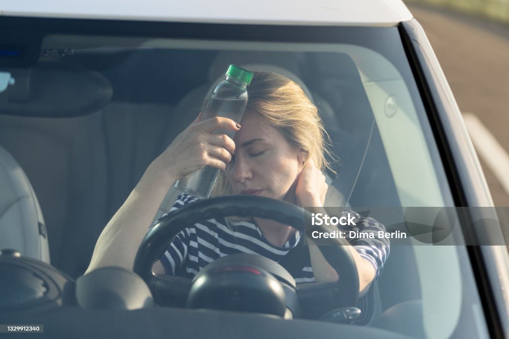 Exhausted girl driver suffering from headache, heat, hot weather applies bottle of water to forehead Exhausted woman driver feeling headache, sitting inside her car, applies bottle of water to forehead, hot weather. Tired female stop after driving car in traffic jam. Blood pressure, heat concept. Car Stock Photo